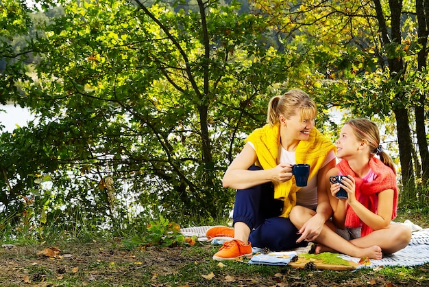 Matka I Córka Bawią Się Na Kocu W Jesiennym Lesie W Pobliżu Rzeki Lub Jeziora. Młoda Matka Bawi Się Z Córką W Jesienny Park W Słoneczny Dzień