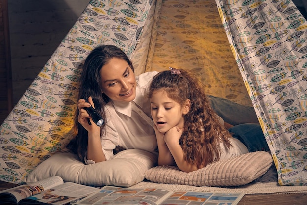 Matka i córka siedzą w namiocie tipi, czytając bajki przy latarce. Szczęśliwa rodzina.