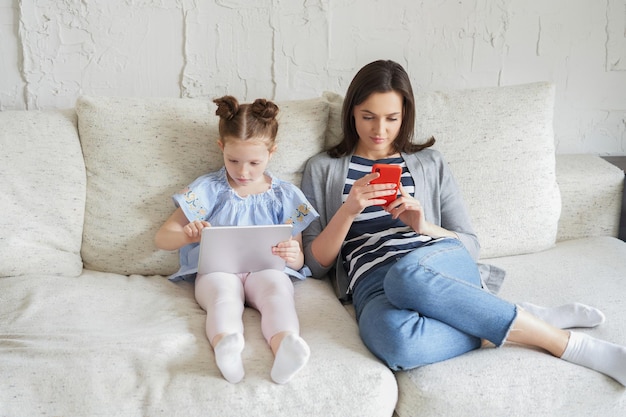 Matka i córka za pomocą urządzeń elektronicznych, siedząc na kanapie w salonie.