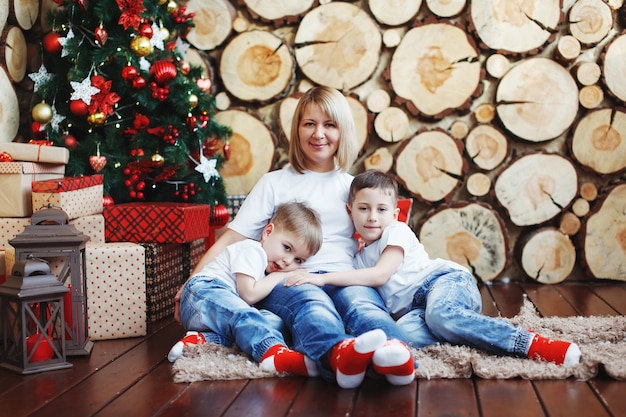 Matka i dwóch chłopców w czapkach świątecznych siedzą przy drzewie noworocznym w tle ściętych drzew.