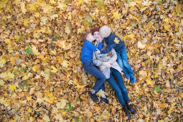 Matka i dwoje dzieci leżących w wielkim stosie liści. Synowie całują mamę. Widok z góry.