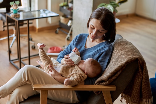 Matka karmi swoje nowo narodzone dziecko z butelki z mlekiem. Młoda europejska kobieta i jej niemowlę dziecko siedzi w drewnianym fotelu. Wnętrze apartamentu typu studio. Pojęcie macierzyństwa i opieki nad dzieckiem