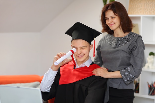 Matka Stoi Za Swoim Synem, Studentem W Sukni Ukończenia Szkoły I Czapce Absolwenta, I Trzyma W Ręku Zwój, A W Domu Ogląda Transmisję Z Ceremonii Ukończenia Szkoły