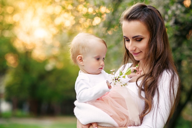 Matka trzyma w ramionach córeczkę wśród kwitnących drzew. Mama i jej małe dziecko nosiły różową rodzinną sukienkę.