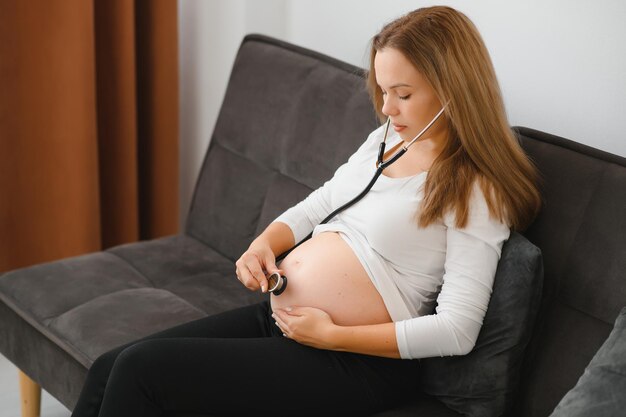 Matka używa stetoskopu, lekarz nasłuchuje odgłosów dziecka w żołądku. Azjatycka kobieta w ciąży za pomocą stetoskopu, słuchając swojego dziecka na kanapie.