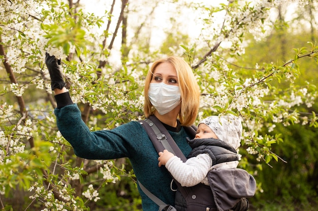 Matka w masce medycznej z małym synkiem w parku spaceruje w pobliżu kwitnącego drzewa