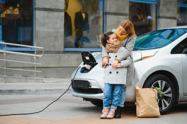 Matka z córką ładującą samochód elektryczny na stacji benzynowej i rozmawiać przez telefon komórkowy.