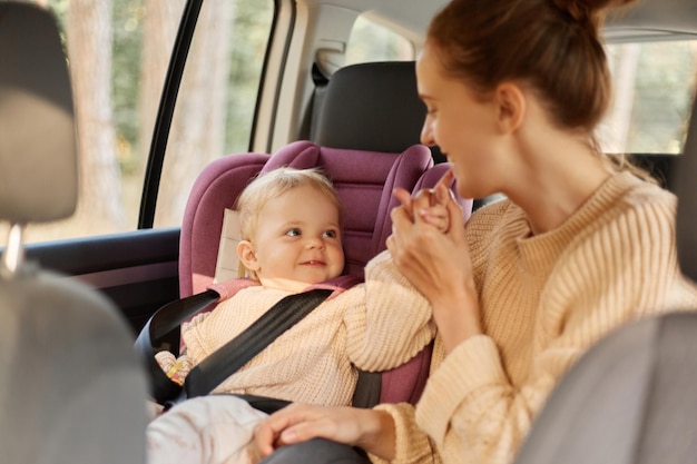 Matka z jej śmieszną córeczką w fotelu bezpieczeństwa w samochodzie, aby jeździć w rodzinnym samochodzie, bawiąc się wokół mamusi, całując lub gryząc palce dziecka lub dziecko wskazujące usta jej mamy