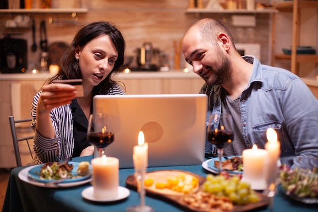 Mąż i żona dokonujący płatności online podczas romantycznej kolacji. Dorośli siedzący przy stole, szukający, przeglądający, surfujący, korzystający z technologii płatności kartą, internet.