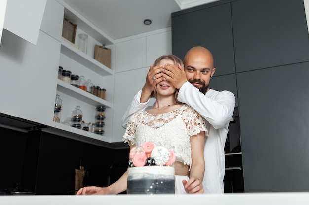 Mąż robi niespodziankę swojej żonie w kuchni, zamykając oczy i przygotowując tort urodzinowy na rocznicową romantyczną koncepcję miłości
