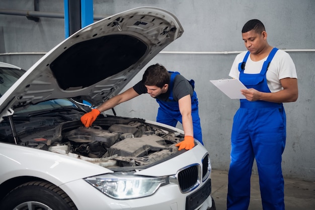 Mechanik W Niebieskim Kombinezonie Bada Zepsuty Silnik Samochodu. Jego Partner Robi Notatki