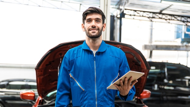 Zdjęcie mechanik w niebieskim mundurze odzieży roboczej, trzymając tablet i patrząc z rozmyciem samochodu w tle.