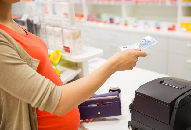 medycyna, farmaceutyka, opieka zdrowotna i koncepcja ludzi - zbliżenie kobiety w ciąży dającej pieniądze i kupującej leki w kasie w aptece