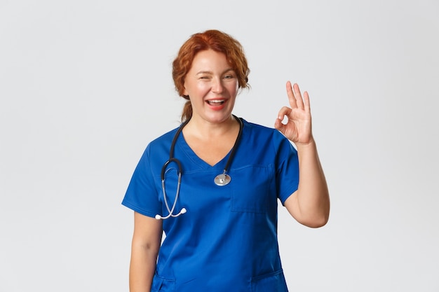Medycyna, koncepcja opieki zdrowotnej. Pewny siebie profesjonalny lekarz zapewnia, że wszystko w porządku, wesoła ruda pielęgniarka w średnim wieku uspokaja pacjenta, pokazuje gest ok i mruga zachęcająco.