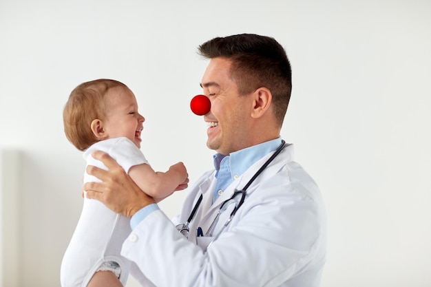 medycyna opieka zdrowotna pediatria i czerwony nos dzień koncepcja szczęśliwy lekarz lub pediatra trzymając dziecko na badaniach medycznych w klinice