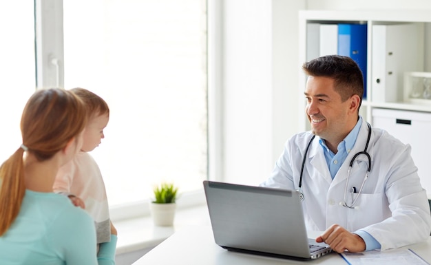 medycyna, opieka zdrowotna, pediatria i koncepcja ludzi - szczęśliwa kobieta z dzieckiem i lekarz z laptopem w klinice