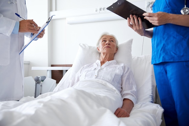 medycyna, wiek, opieka zdrowotna i koncepcja ludzi - lekarz i pielęgniarka ze schowkami odwiedzająca starszą pacjentkę na oddziale szpitalnym