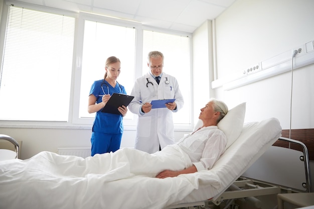Zdjęcie medycyna, wiek, opieka zdrowotna i koncepcja ludzi - lekarz i pielęgniarka ze schowkami odwiedzająca starszą pacjentkę na oddziale szpitalnym