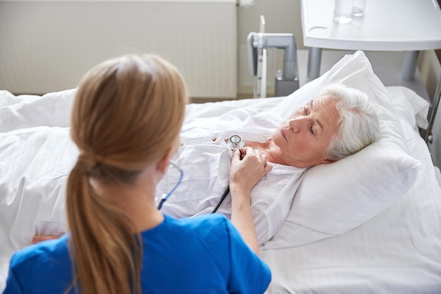 medycyna, wiek, wsparcie, opieka zdrowotna i koncepcja ludzi - lekarz lub pielęgniarka ze stetoskopem odwiedza starszą kobietę i sprawdza jej oddech lub bicie serca na oddziale szpitalnym