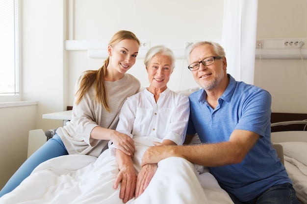 medycyna, wsparcie, rodzinna opieka zdrowotna i koncepcja ludzi - szczęśliwy starszy mężczyzna i młoda kobieta odwiedzający i dopingujący babcię leżącą w łóżku na oddziale szpitalnym
