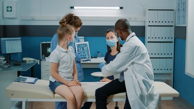 Medyk i pielęgniarka wyjaśniające wyniki skanowania rentgenowskiego na tablecie w szafce. Lekarz ogólny i asystent pokazujący obraz radiologiczny na ekranie małej dziewczynce i matce podczas badania kontrolnego.
