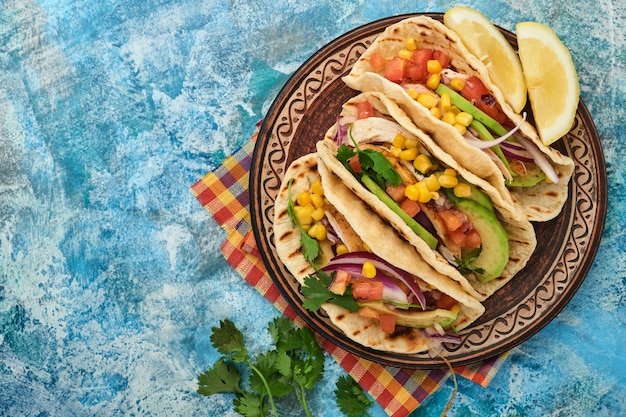 Meksykańskie tacos z grillowanym kurczakiem, awokado, ziarnami kukurydzy, pomidorem, cebulą, kolendrą i salsą przy niebieskim kamiennym stole. Tradycyjne meksykańskie i latynoamerykańskie jedzenie uliczne. Widok z góry.