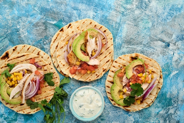 Meksykańskie tacos z grillowanym kurczakiem, awokado, ziarnami kukurydzy, pomidorem, cebulą, kolendrą i salsą przy niebieskim kamiennym stole. Tradycyjne meksykańskie i latynoamerykańskie jedzenie uliczne. Widok z góry.