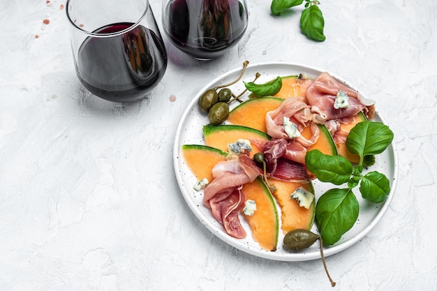 Melon kantalupa z szynką prosciutto lub jamon i zielonymi liśćmi bazylii z lampką wina pyszna sałatka superfood na jasnym tle Tradycyjna włoska przekąska antipasti miejsce na tekst widok z góry