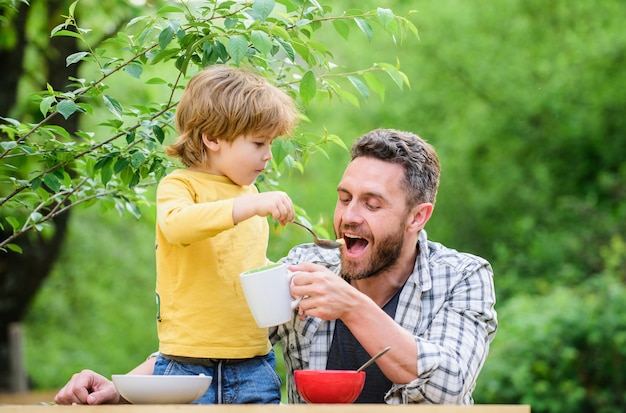 Menu dla dzieci. Rodzina cieszy się domowym posiłkiem. Nawyki żywieniowe. Mały chłopiec z tatą jedzenie natura tło. Letnie śniadanie. Koncepcja zdrowej żywności. Ojciec syn je jedzenie i baw się dobrze. Karmienie dziecka.