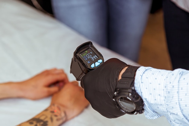 Męska dłoń w czarnej rękawiczce mierzy temperaturę na ramieniu pacjenta.