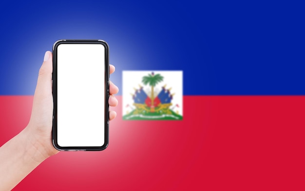 Męska ręka trzymająca smartfon z pustym ekranem na tle niewyraźnej flagi Haiti Widok zbliżenia
