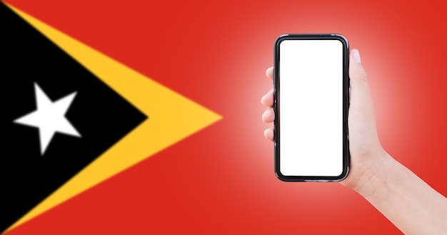 Męska ręka trzymająca smartfon z pustym ekranem na tle niewyraźnej flagi Timoru Wschodniego Widok zbliżenia