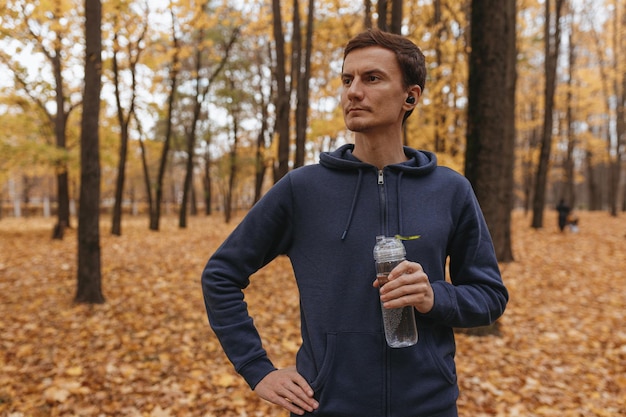 Męski biegacz z butelką wody wielokrotnego użytku stojącą w jesiennym parku i robiącą sobie przerwę podczas treningu