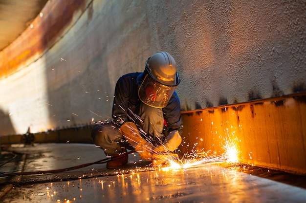 Męski pracownik iskra do cięcia metalu na stalowej płycie dna zbiornika z błyskiem światła tnącego z bliska nosić rękawice ochronne i maskę w bocznej ograniczonej przestrzeni.