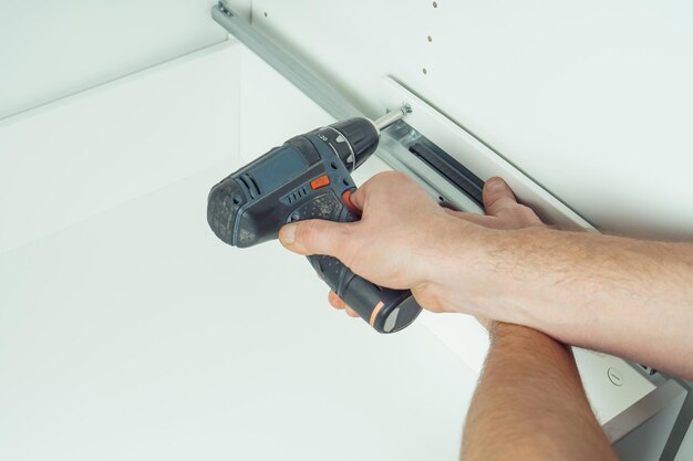 Męskie dłonie trzymają śrubokręt elektryczny akumulatorowy, aby naprawić szufladę montażową szyny białej komody lub szafy