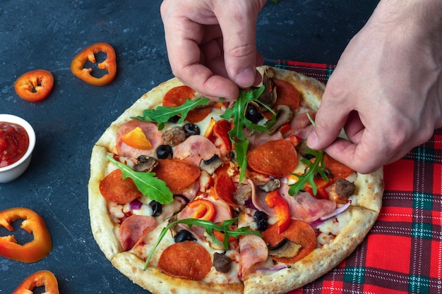 Męskie dłonie zdobią pizzę zielenią. Świeża przygotowana pizza z salami, pieczarkami, szynką i serem na ciemnym tle. Tradycyjny włoski obiad lub kolacja. Ścieśniać