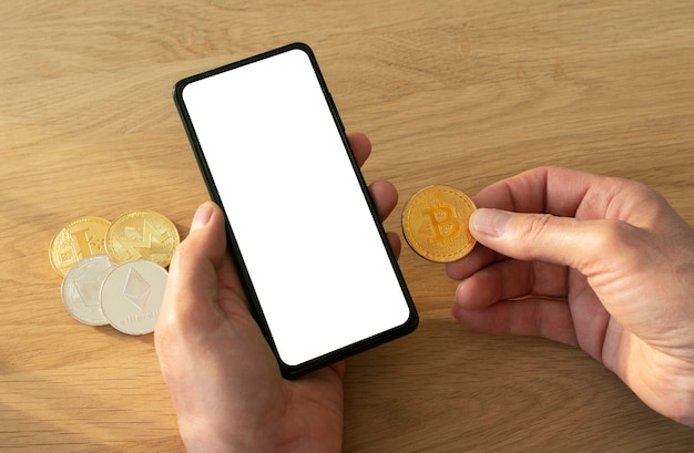 Męskie ręce trzymając telefon komórkowy z ekranem do makiety i monety bitcoin w ręku na drewnianym stole.