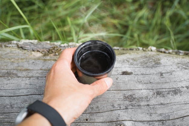 Męskiej ręki trzymającej kubek gorącej herbaty na zewnątrz w lesie, zbliżenie. Para unosi się z napoju w stalowym kubku na kłodzie na zewnątrz. Koncepcja kempingu