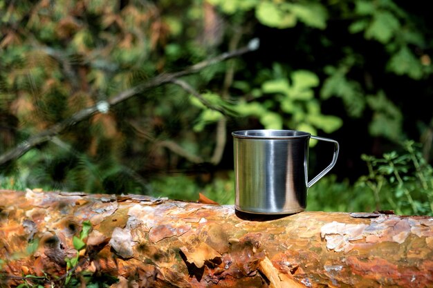Metalowy kubek turystyczny z gorącym napojem stoi na pniu drzewa w lesie Turystyka na świeżym powietrzu