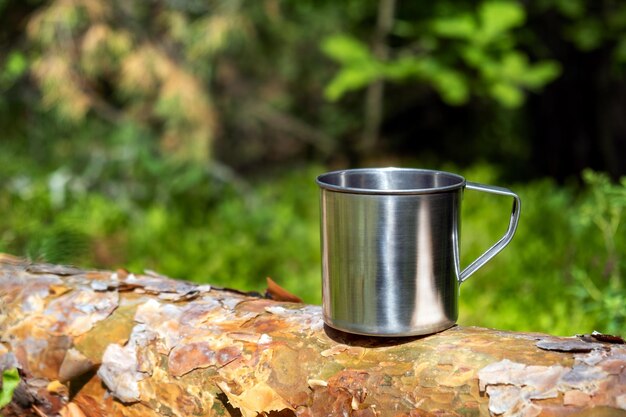 Metalowy kubek turystyczny z gorącym napojem stoi na pniu drzewa w lesie