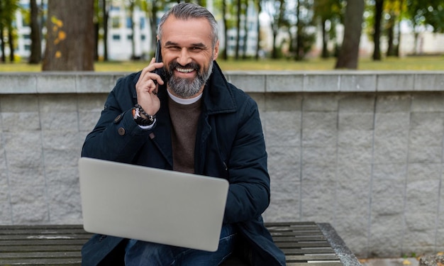 Mężczyzna biznesmen pracujący w parku laptopa rozmawia przez telefon uśmiechając się patrząc na kamerę