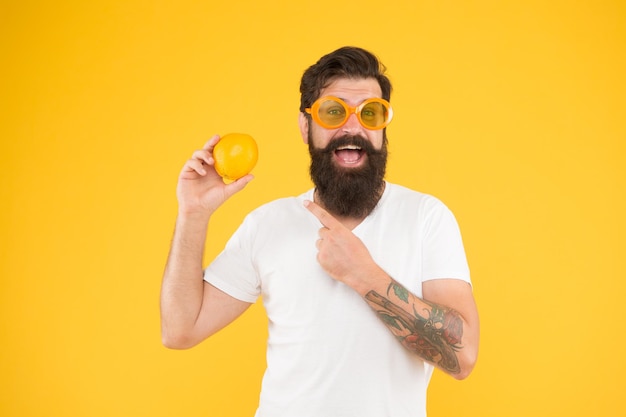 Mężczyzna brodaty hipster w pomarańczowych okularach przeciwsłonecznych na żółtym tle Wesoły facet trzyma dojrzałe pomarańczowe owoce cytrusowe Letnie wakacje Źródło orzeźwienia Letnie odżywianie Hipster z brodą w letnim nastroju