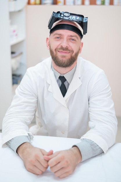 Mężczyzna brodaty lekarz w płaszczu medycznym iz powiększającym paskiem na głowę ze światłami siedzącymi na biurku Sprzęt optyka Gabinet medyczny
