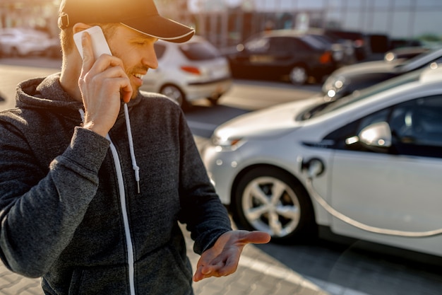 Mężczyzna Czeka, Aż Jego Samochód Elektryczny Będzie ładował ładowarkę Na Parkingu W Pobliżu Centrum Handlowego.