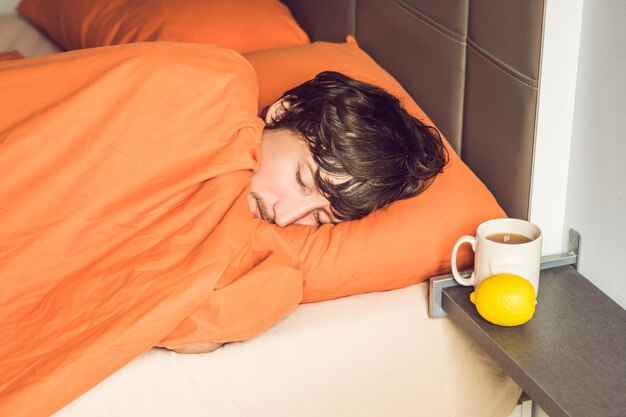 Mężczyzna czuje się zimno, leży w łóżku i pije herbatę i mierzy temperaturę termometrem
