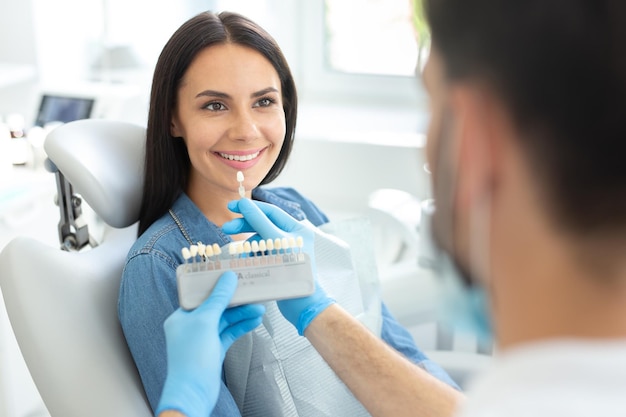 Mężczyzna dentysta trzyma paletę kolorów zębów, podczas gdy piękna uśmiechnięta kobieta patrzy na niego w klinice