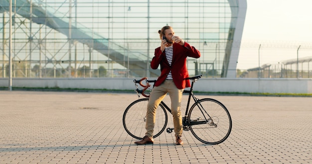 Mężczyzna dorywczo przystojny rowerzysta w czerwonej kurtce siedzi na rowerze, sącząc gorący napój i rozmawiając przez telefon. W dużym przeszklonym nowoczesnym budynku. Kaukaski mężczyzna pije kawę. i rozmawiając przez telefon komórkowy.