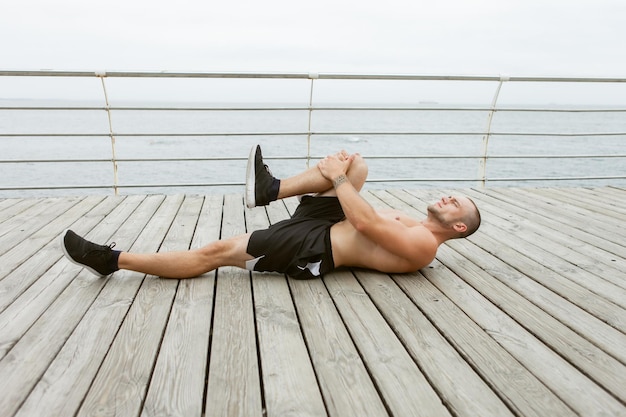 Mężczyzna fitness z nagim torsem robi rozciąganie nóg leżąc na drewnianej desce na plaży