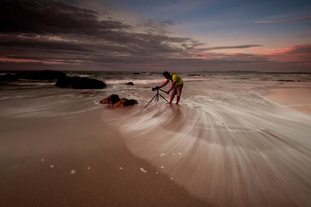 Mężczyzna fotografuje na brzegu na plaży przeciwko niebu podczas zachodu słońca