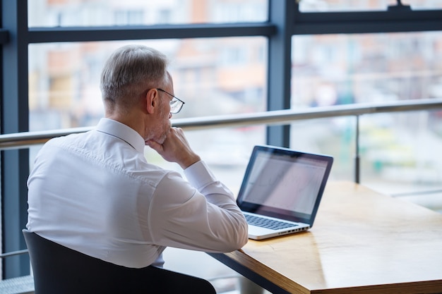 Mężczyzna freelancer pracuje w kawiarni nad nowym projektem biznesowym. Siedzi przy dużym oknie przy stole. Patrzy na ekran laptopa z filiżanką kawy